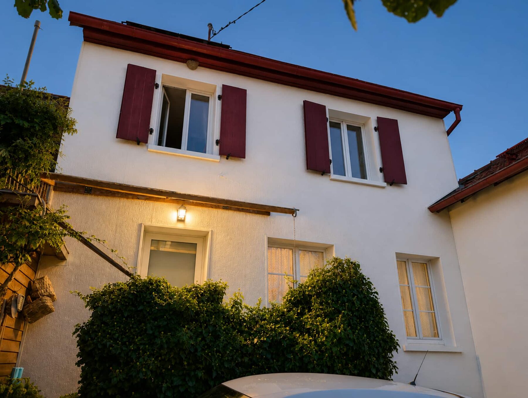 La façade de la maison au crépuscule : volets rouges, lanterne allumée et plantes grimpantes